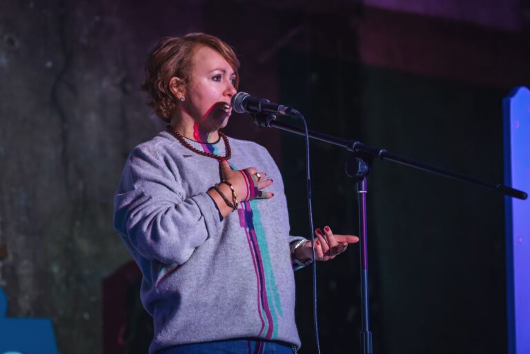 Cat Cochrane performs at the Scottish National Poetry Slam Championship at the Panopticon in Glasgow on Feb 14. Cat stands at the mic looking out to the audience. Cat has red hair and is wearing a grey jumper.