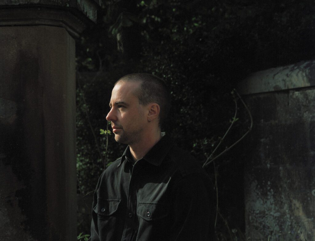 Upper body image of Len Lukowski. Len is wearing black shirt stood outside gates of cemetery, with evening sun on his face and. Foliage and stone visible in background.