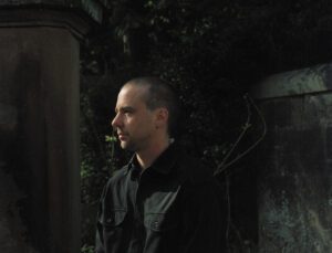 Upper body image of Len Lukowski. Len is wearing black shirt stood outside gates of cemetery, with evening sun on his face and. Foliage and stone visible in background.