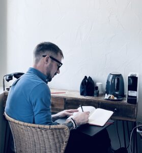 The poet sits in a hotel room, facing away from the camera, writing his diary.