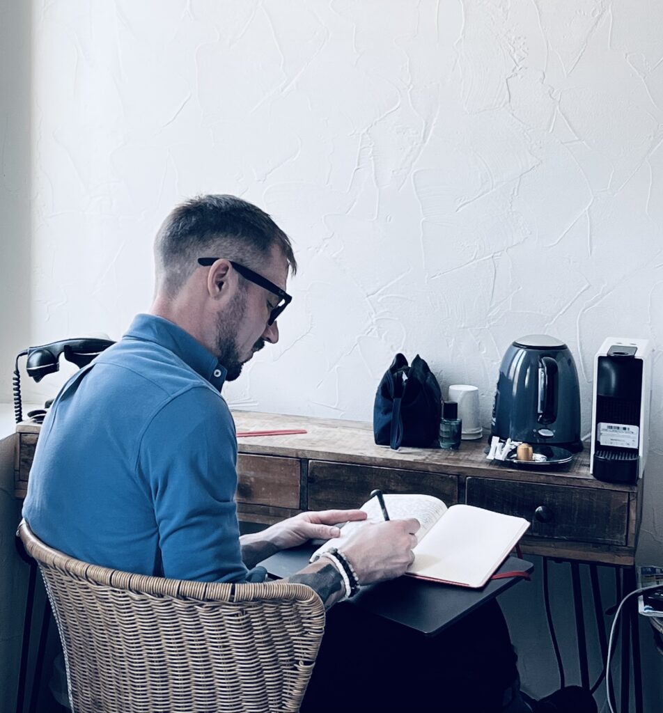 The poet sits in a hotel room, facing away from the camera, writing his diary.