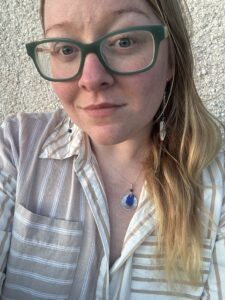 Photo of Sam wearing her handmade coastal jewellery. A cobalt blue sea glass sterling silver necklace paired with blue and white mismatched pottery earrings. Sam has long ginger hair, freckles, blue eyes and wears teal coloured glasses with a brown and white striped cotton shirt.