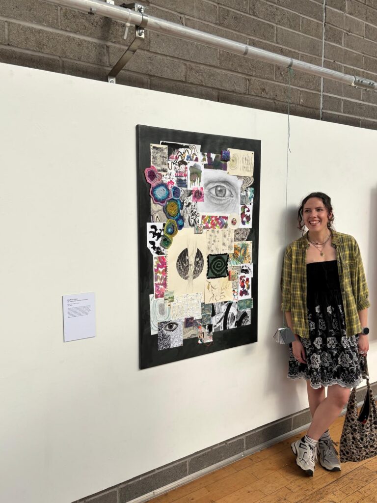 Lily standing next to her art piece at the exhibition "the art of becoming" in her final year of studying art psychotherapy at queen margaret university. she is smiling and looking off to the side. her art piece is a large collage of many smaller artworks.