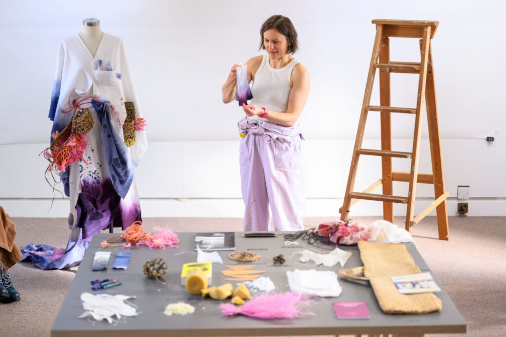 A woman stands in a white, bright studio. Behind her is a costume she's in the process of making, that looks a bit like a kimono. There is also a wooden stepladder. In front of her is a table with bits of materials on it - rubbish, textiles, and mystery objects.