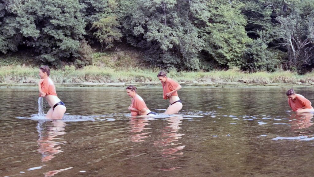 A group of women in orange t-shirts and black bikini bottoms running happily through a river.