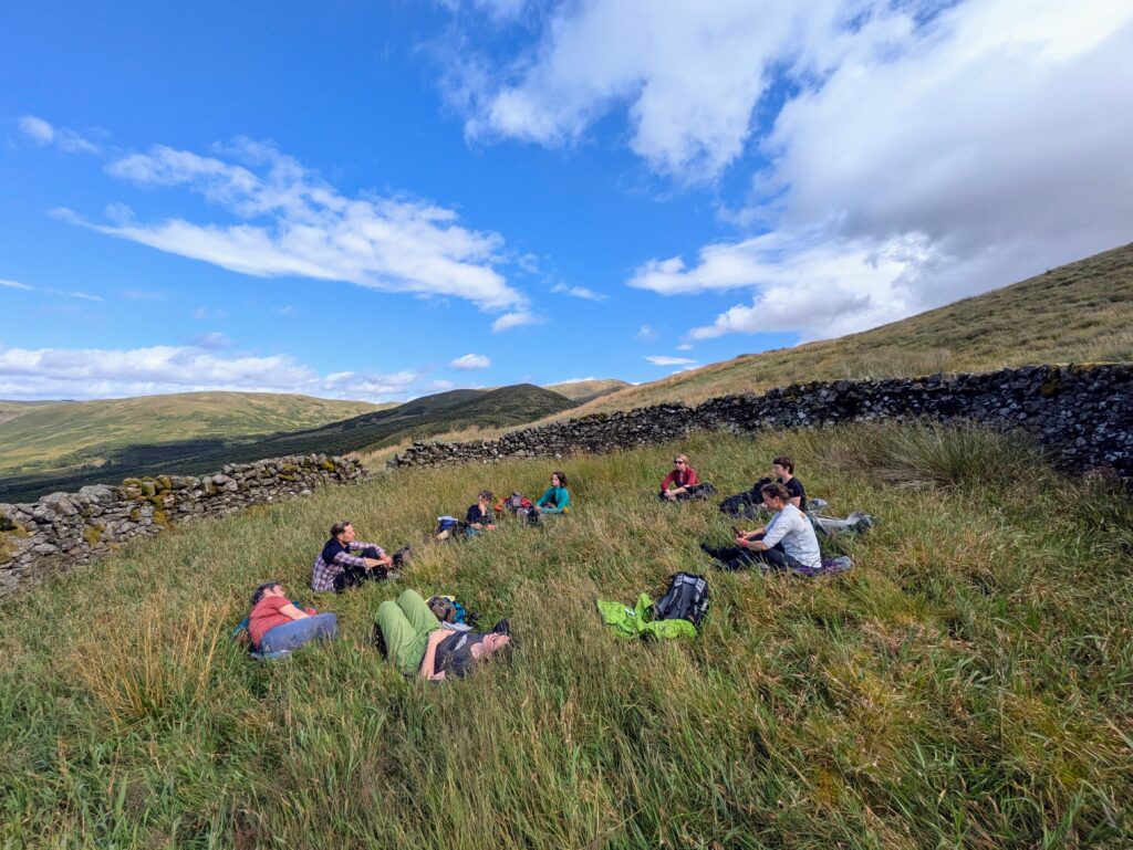 A group of people sitting in a circle, in long grass. Above them is a blue sky with fluffy white clouds.