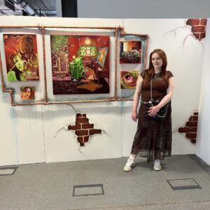 This is me at my degree show, standing beside my final installation work consisting of five paintings in different sizes surrounded by sculpted copper pipes on a background with verdigris and patches of exposed brick, which I also painted.  