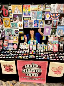 A colourful art stall displaying illustrated prints, stickers, badges, and zines. Chloe sits at the centre in a blue top, behind the table, surrounded by artwork featuring cartoon characters, animals, and bold designs. A pink banner sits across the front, advertising Coin-Operated Press.